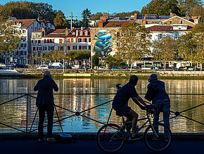 Au premier plan des photographes et un homme à vélo, au second plan l'Adour. De l'autre côté du fleuve au loin sur tout un pan de mur sont peintes dans un camaieu de bleu des baleines posées les unes sur les autres. - Irudia handitu (modu-leihoa)