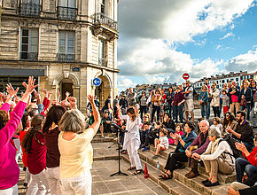 Sur la gauche un groupe de chanteurs, les bras levés chantent en suivant les instructions de leur chef d'orchestre devant de nombreux spectateurs par beau temps. - Irudia handitu (modu-leihoa)
